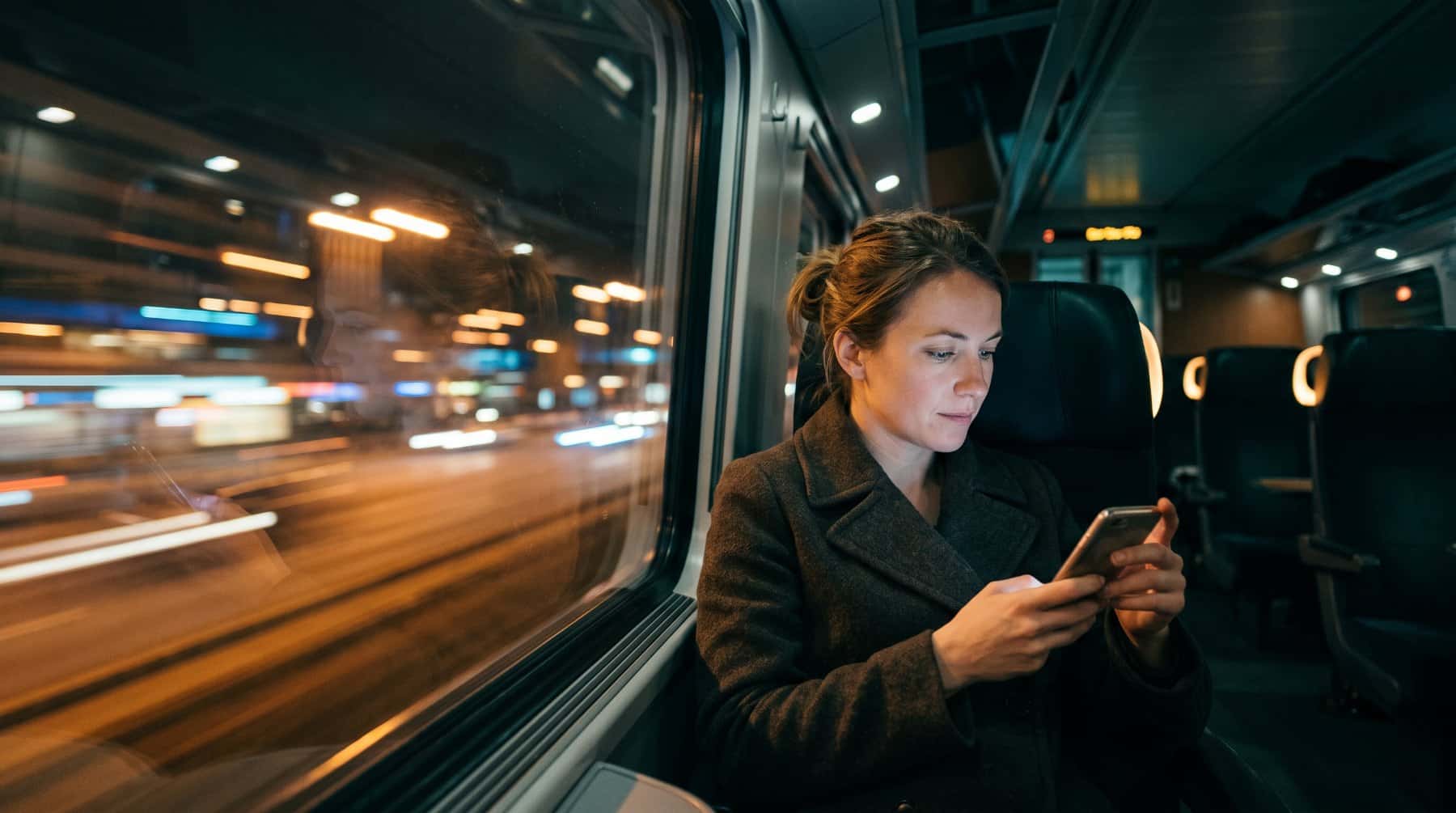 Passenger using a phone on a night train as city lights blur past the window